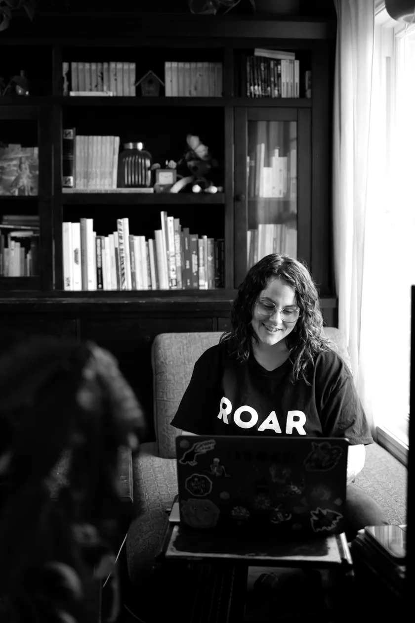 Photo en noir et blanc de Stéphanie qui sourit en travaillant à l’ordinateur, devant une bibliothèque.