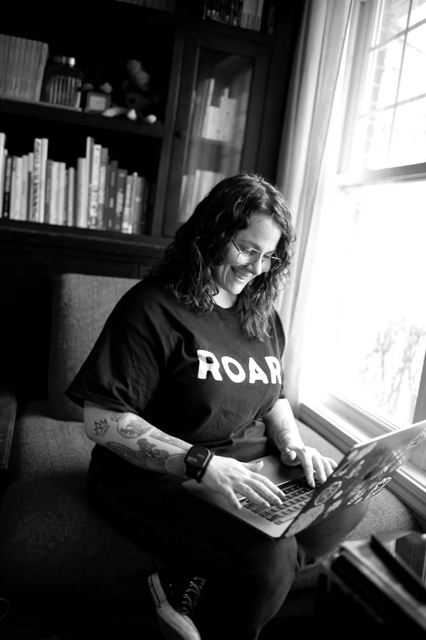 Photo en noir et blanc de Stéphanie qui tape à l’ordinateur, assise près d’une fenêtre, en t-shirt ROAR.
