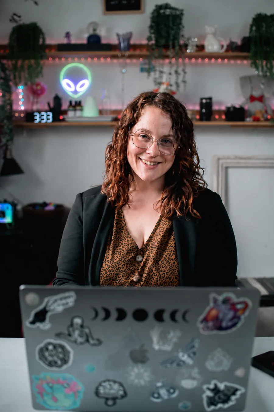 Stéphanie Guilbeault souriante derrière son ordinateur couvert d’autocollants, devant des étagères avec plantes et néon d’extraterrestre.
